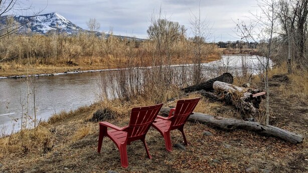 Two red Adirondack chairs by a river with a mountain in the background.