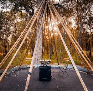 Estructura de madera con un banco y una parrilla portátil en un bosque.