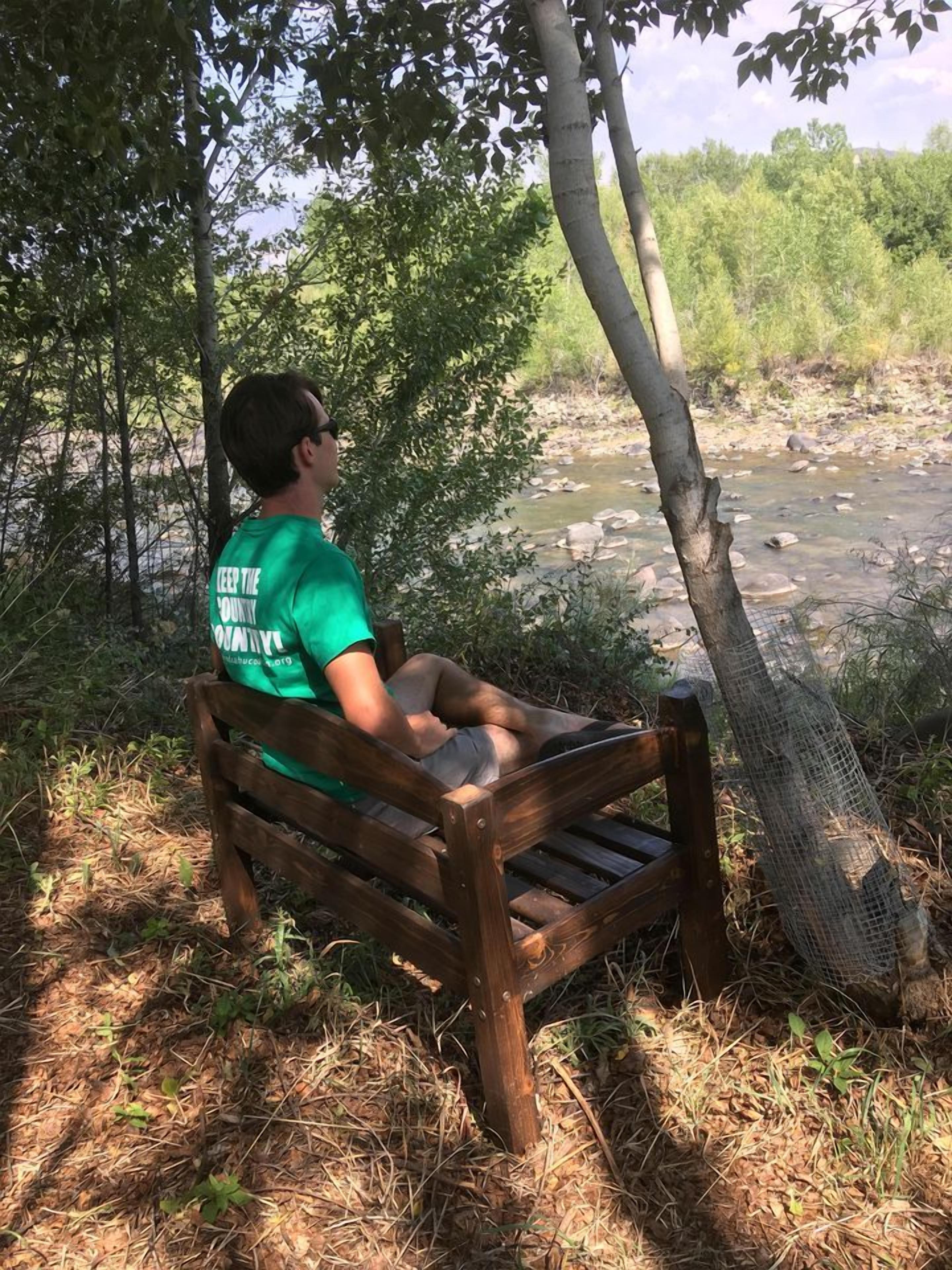 Iemand zit op een houten bankje in een natuurlijke omgeving, met een rivier en bomen op de achtergrond.