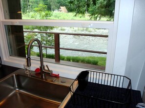 A kitchen with a stainless steel sink, a window with a view of a river and greenery, and a black metal rack.