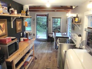 A compact kitchen with a microwave, toaster, and refrigerator. There is a dining area with a chair and a table. The room has wooden flooring and a large window. - Pet-Friendly Cabin Rental Overlooking a River near Skykomish, Washington (Baring)