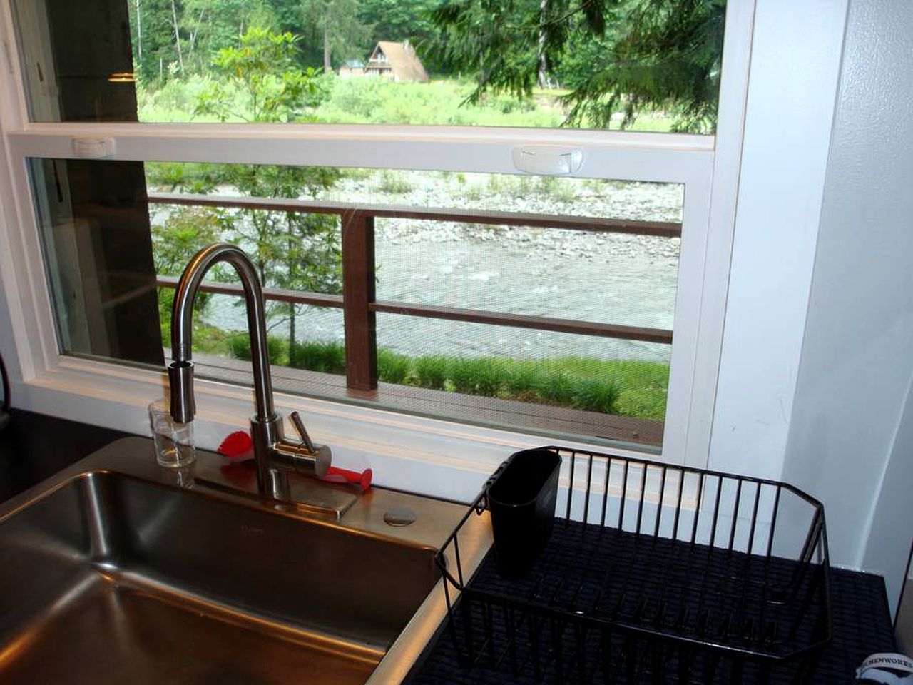 A kitchen with a stainless steel sink, a window with a view of a river and greenery, and a black metal rack.