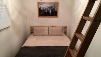 A small bedroom with a bed, a wooden ladder, a framed picture on the wall, and a window with blinds.