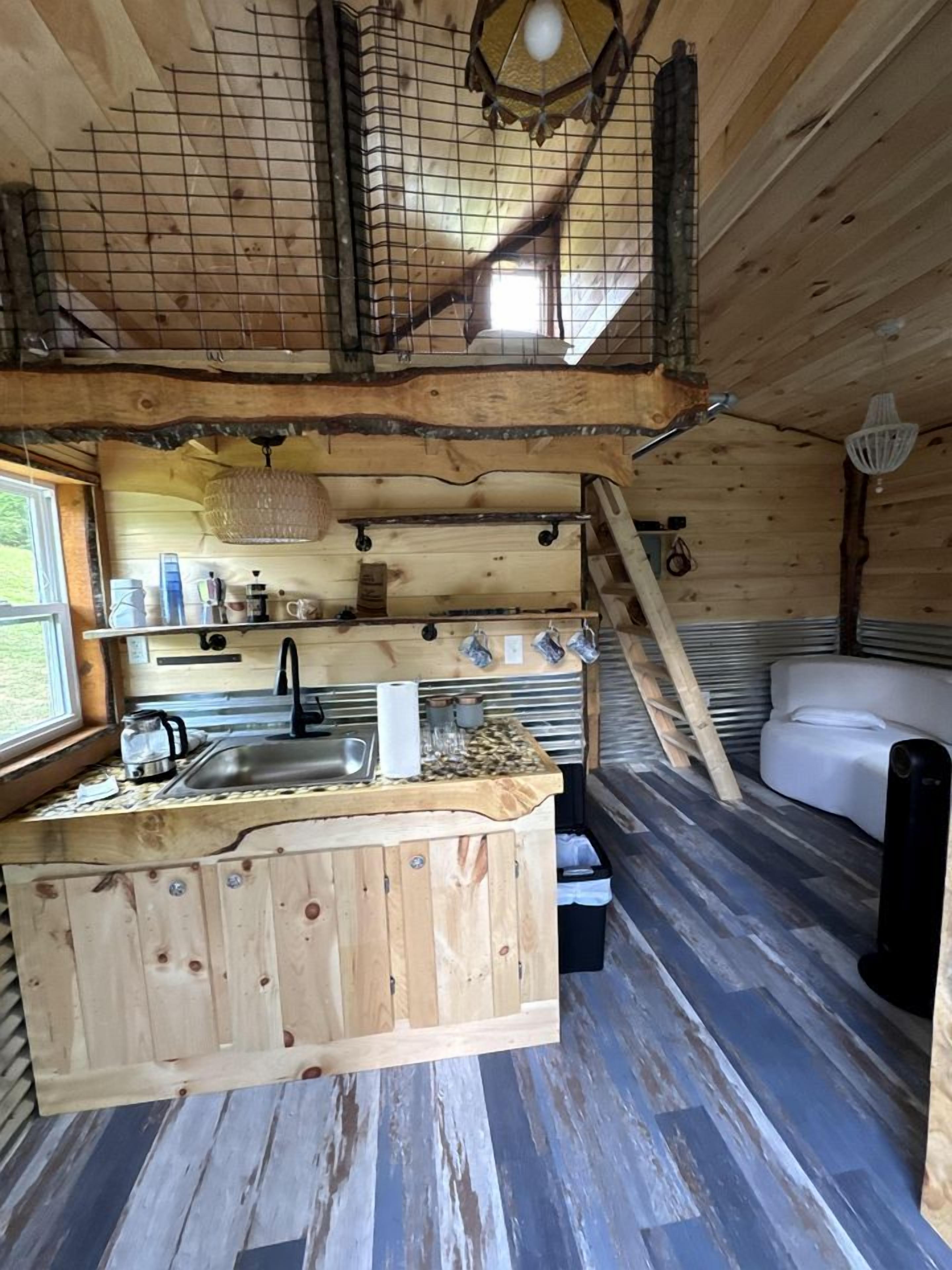 A cabin interior with a wooden floor, a kitchen area with a sink, a white couch, and a ladder leading to a loft space.
