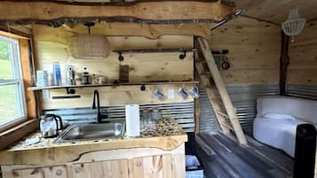A cabin interior with a wooden floor, a kitchen area with a sink, a white couch, and a ladder leading to a loft space.