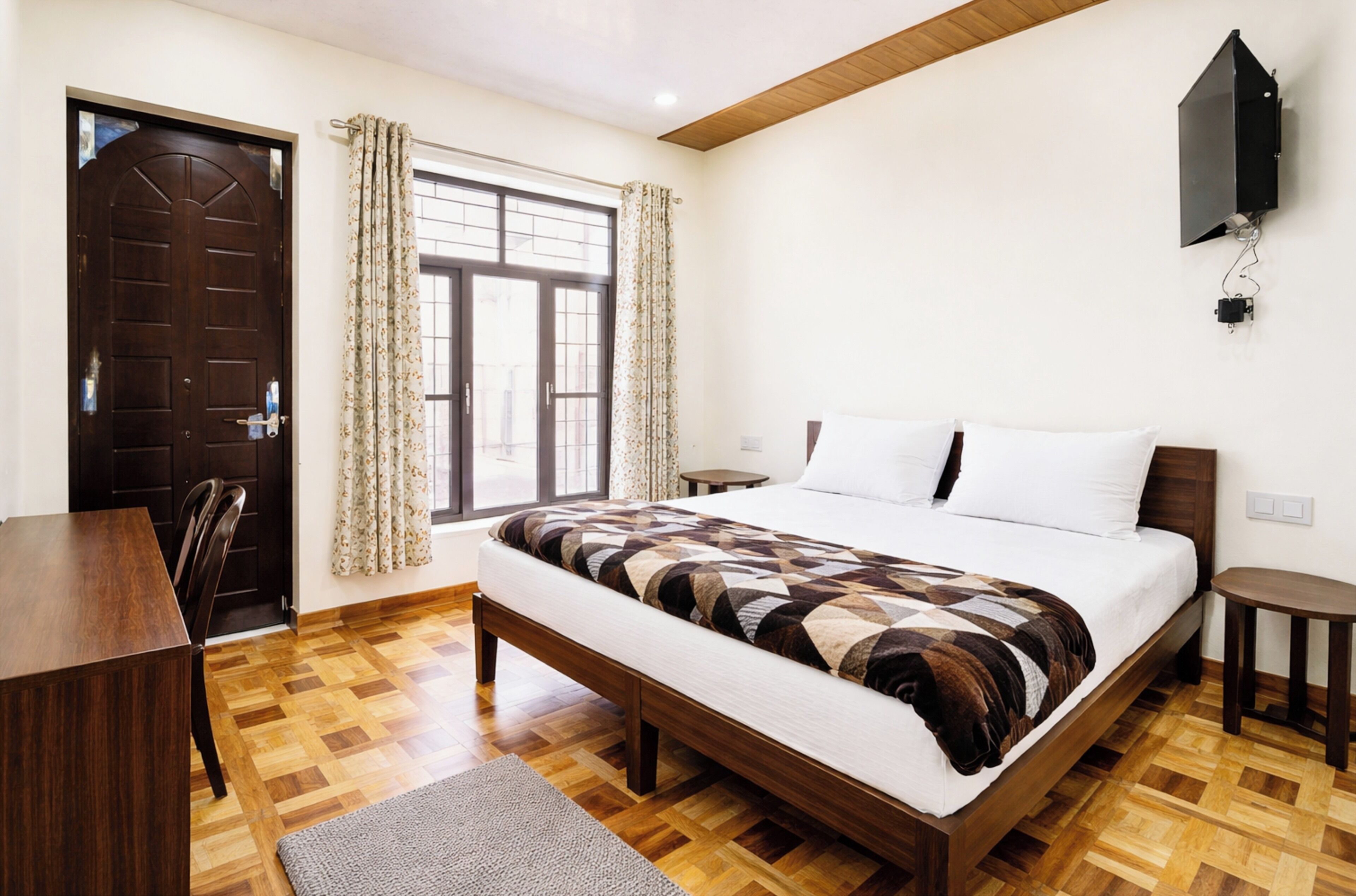 A bedroom with a wooden bed, a desk, a chair, a window with curtains, and a mounted TV.