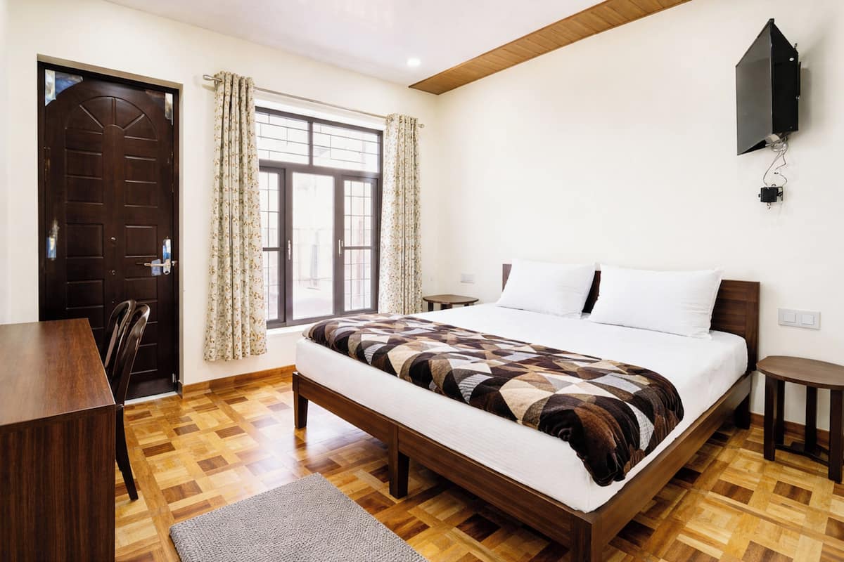 Un dormitorio con cama de madera, un escritorio, una silla, un ventanal con cortinas y un televisor montado.