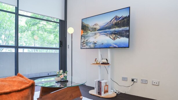 Een moderne woonkamer met een groot raam, een flatscreen televisie aan de muur, een glazen salontafel en een decoratieve plank met boeken en een beeldje.