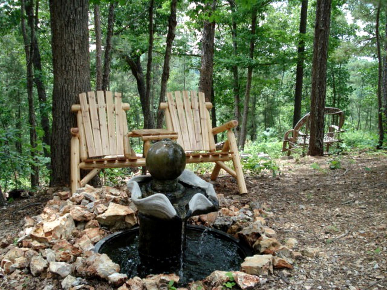 Un cadre extérieur rustique agrémenté d’une fontaine en pierre, de deux fauteuils Adirondack en bois et d’un paysage boisé en arrière-plan.