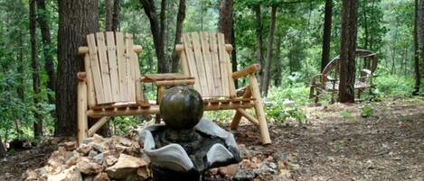Un entorno rústico al aire libre con una fuente de piedra, dos sillas de madera tipo Adirondack y un fondo de bosque.