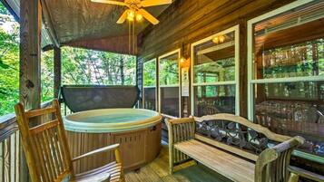 A wooden deck with a hot tub, wooden chairs, a bench, and large windows overlooking a forest.