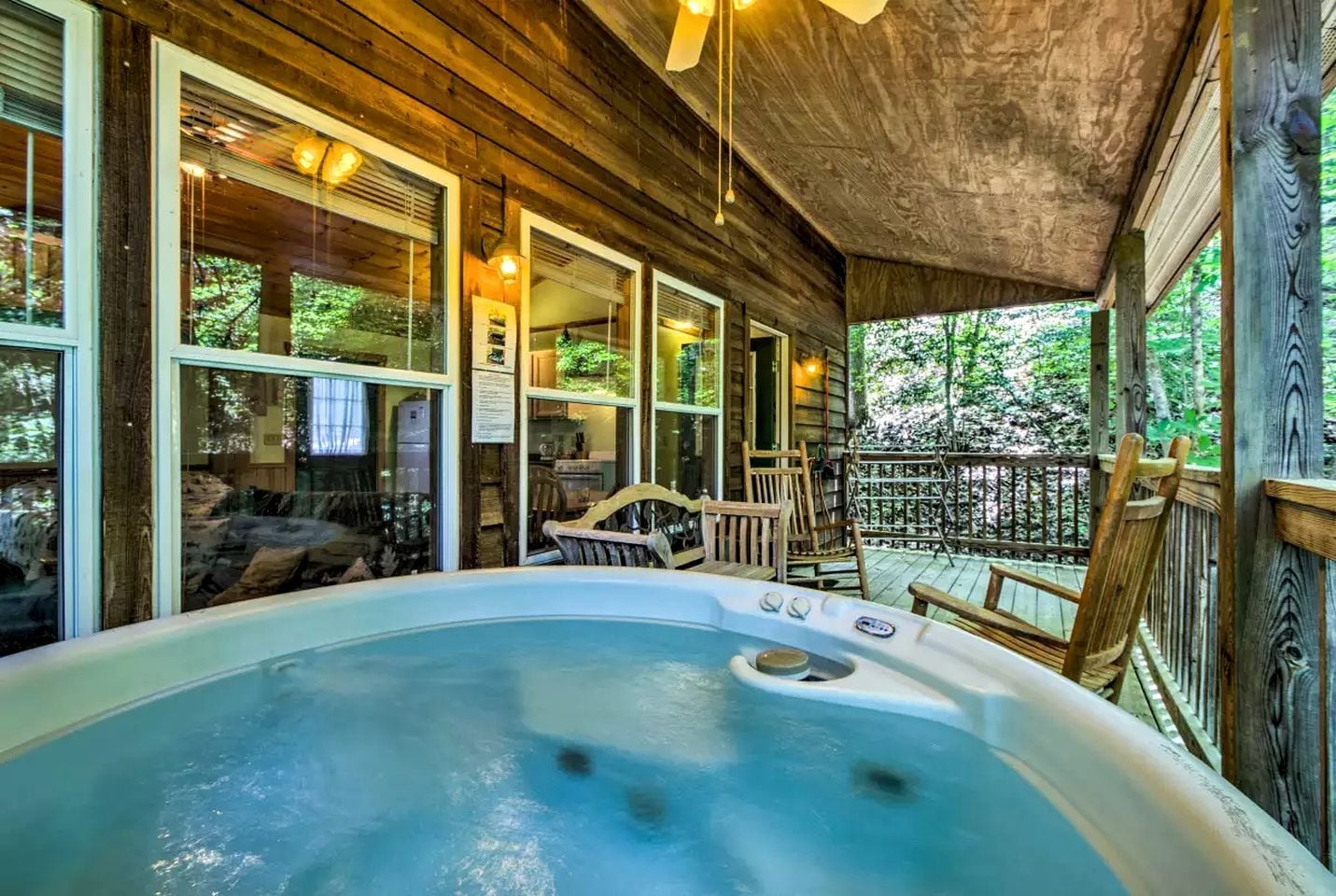 A hot tub in a wooden cabin with a covered porch, large windows, and a view of the surrounding forest.