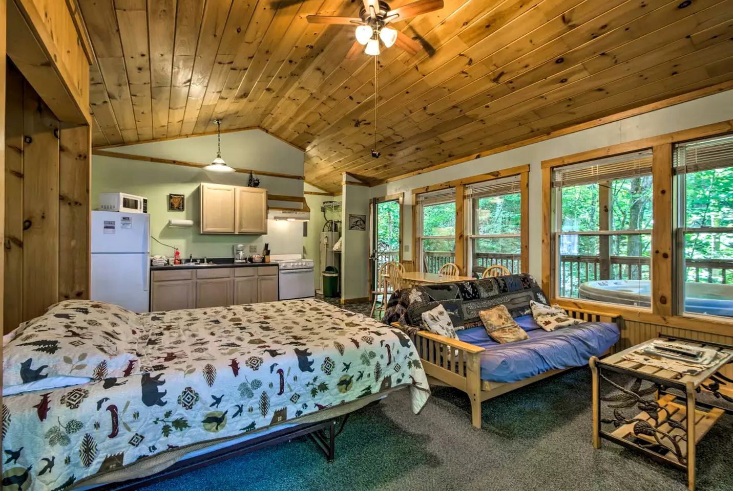 Une cabane avec un intérieur en bois, une kitchenette, un lit avec une literie à motifs, un canapé avec des coussins et de grandes fenêtres.