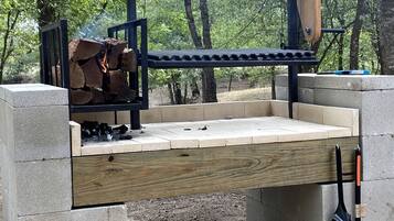 A concrete outdoor grill with a wooden bench, surrounded by trees.