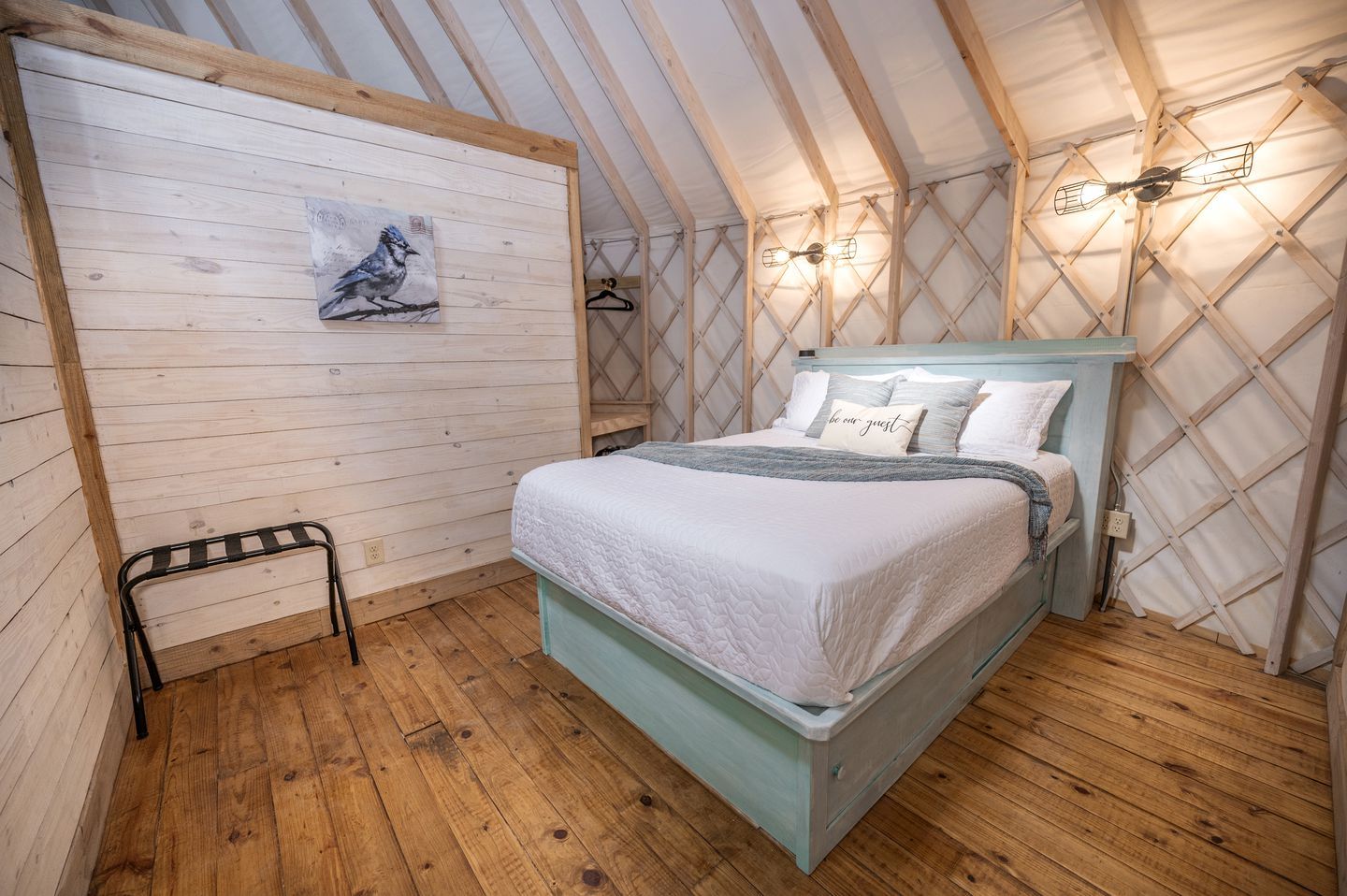 Una habitación acogedora con cama individual, piso de madera, cabecera de cama azul claro, un cuadro en la pared y una lámpara colgante.