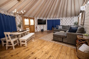 A tented accommodation with a wooden floor, a sofa, a dining table with chairs, and a TV mounted on the wall.