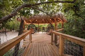 Eine Holzterrasse mit einem Gazebo mit Reetdach, umgeben von Bäumen und ausgestattet mit Korbstühlen und einer Hängeschaukel.