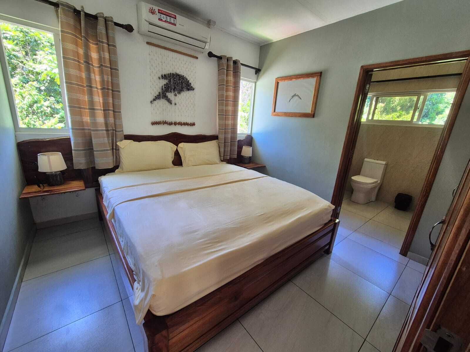Una habitación de hotel con una cama, mesitas de noche, un equipo de aire acondicionado, un cuadro enmarcado en la pared y la puerta del baño.