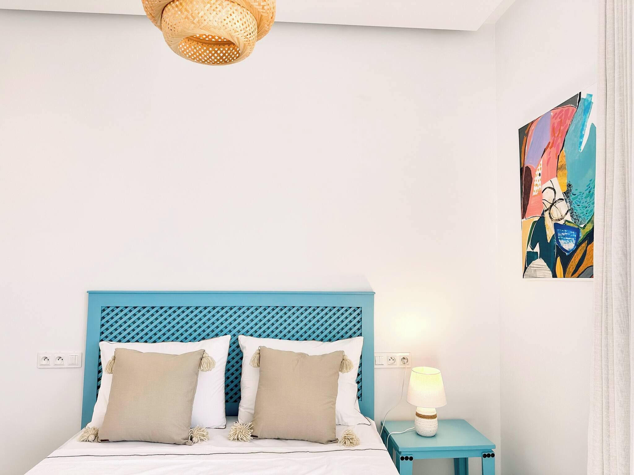 A bedroom with a blue headboard, two pillows, a bedside lamp, and a painting on the wall.