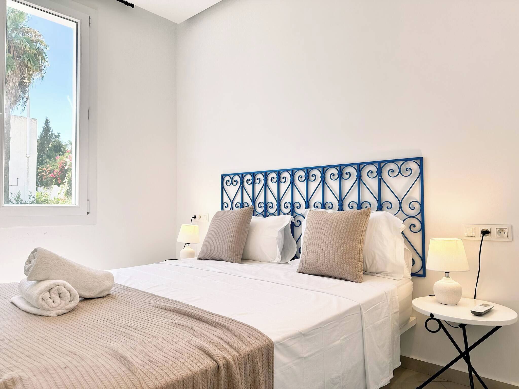 A bedroom with a bed, two bedside tables with lamps, a window with a view of greenery, and a decorative metal headboard.