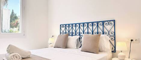A bedroom with a bed, two bedside tables with lamps, a window with a view of greenery, and a decorative metal headboard.