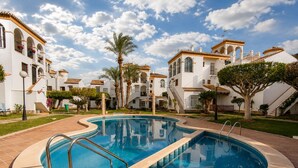 A swimming pool with a curved design, surrounded by a paved walkway, and adjacent to a building with multiple balconies and a tiled roof. - Ground Floor in Playa Flamenca with Pool (Alicante)