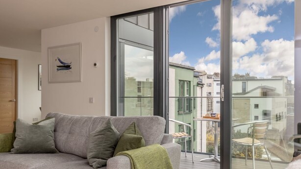 A modern living room with a grey sofa, a green throw blanket, and a large sliding glass door leading to an outdoor balcony. - Designer Urban Apartment with City Terrace (Edinburgh)