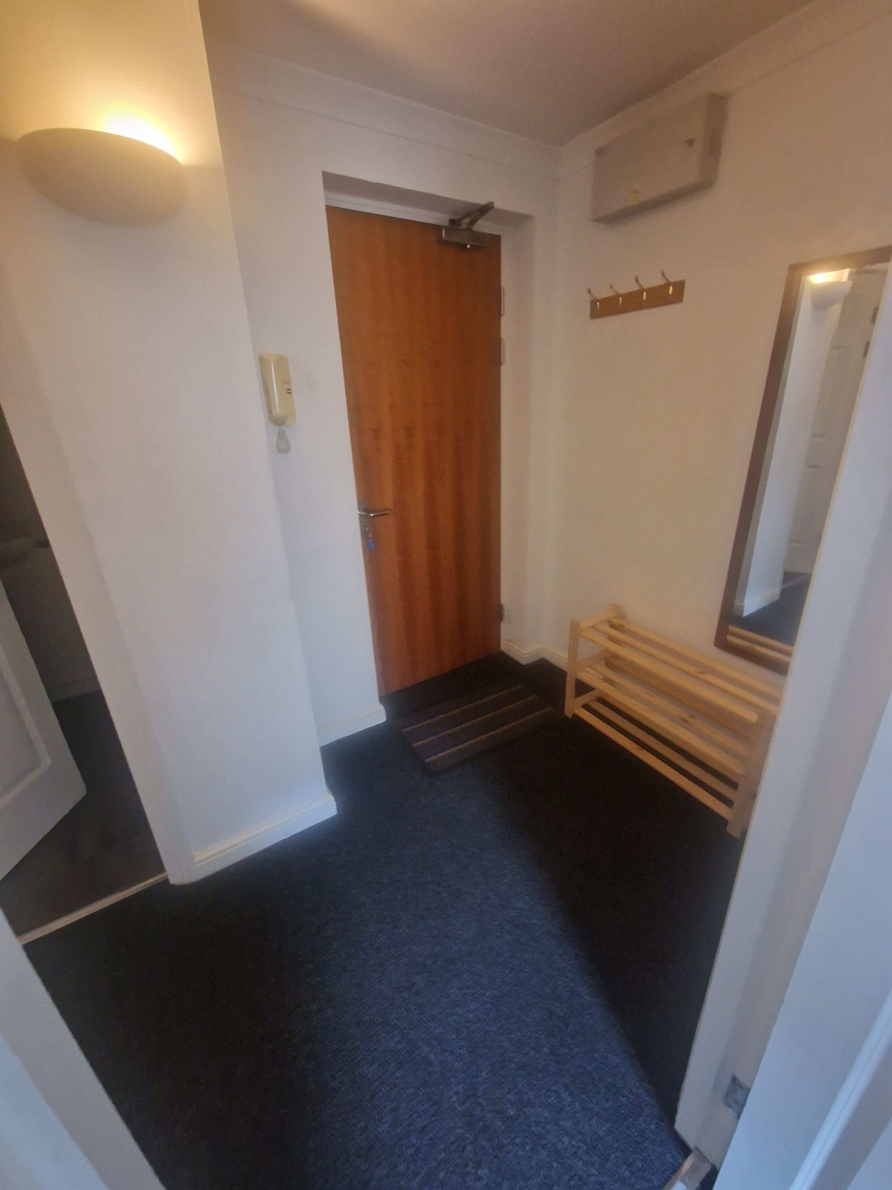 Un pasillo con una puerta de madera, una lámpara de pared, un espejo y un banco de madera.