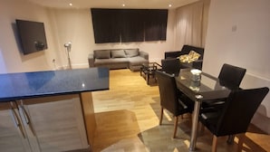 Een moderne woonkamer met een bank, een eettafel met stoelen, een keuken met een aanrecht en een wandgemonteerde flatscreen-tv.
