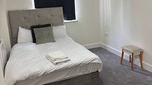 Een net opgemaakt bed met witte lakens, een grijze hoofdbord en een klein houten krukje.