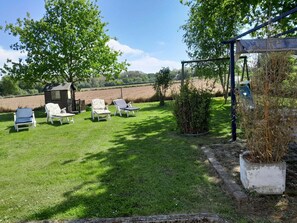 Un espacio exterior con césped, cuatro sillas de lounge blancas, una pequeña estructura de madera y un jardín con plantas.