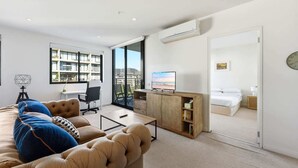 Un salon moderne avec un canapé, un bureau et un téléviseur. On aperçoit une chambre à coucher avec un lit et une salle de bain à travers une porte ouverte.