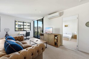 Ein modernes Wohnzimmer mit Sofa, Schreibtisch und Fernseher. Man sieht durch eine geöffnete Tür außerdem ein Schlafzimmer mit Bett und ein Badezimmer.