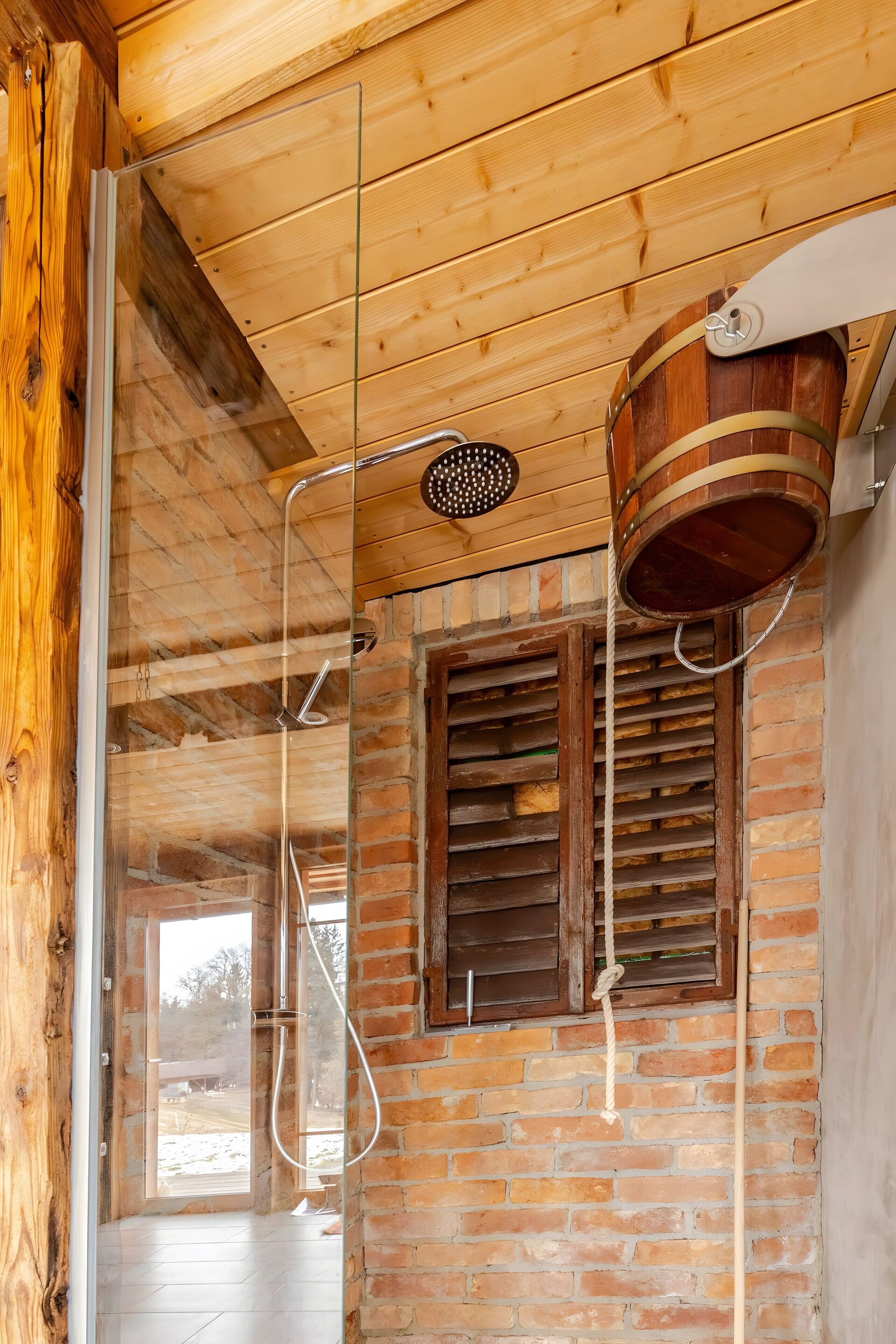 Un plafond en bois, une porte en verre, un espace douche avec un tuyau d’arrosage et un seau en bois.