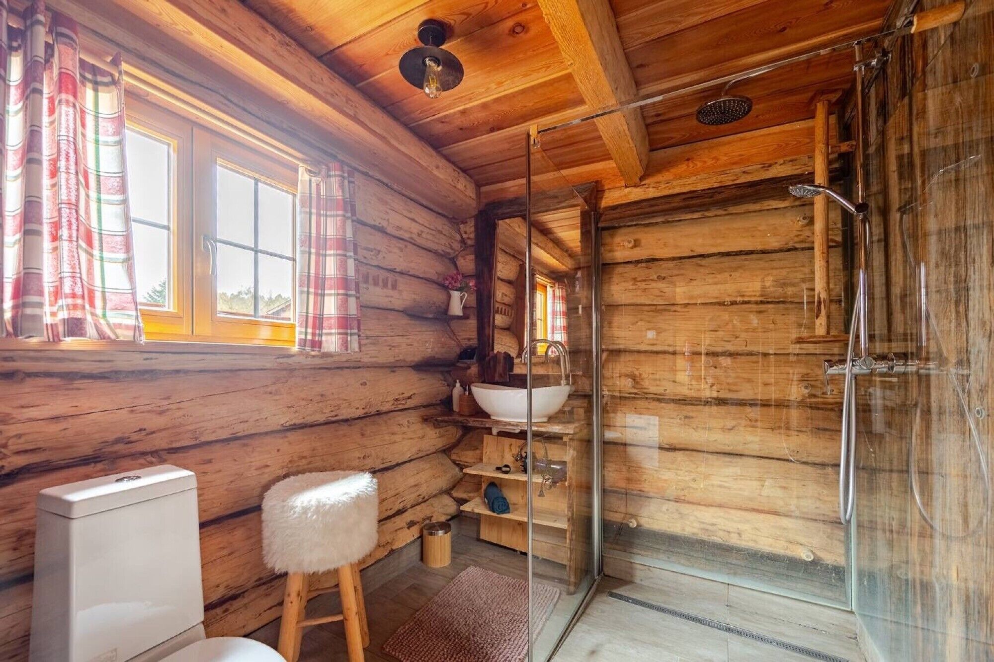 Ein Blockhäuschen-Interieur mit Holzvertäfelung, einer Duschkabine aus Glas, einem weißen Waschbecken und einem Holzschemel.