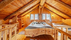 Uma cabana de madeira com um quarto no mezanino, que possui uma cama, cabeceira de madeira, uma janela e um banco de madeira.
