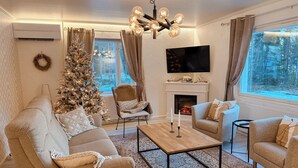 Uma sala de estar acolhedora com uma árvore de Natal, lareira, televisão e uma janela grande com vista para árvores.