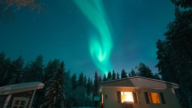 Uma cabana pequena com telhado coberto de neve, cercada por árvores, sob um céu noturno com a aurora boreal.