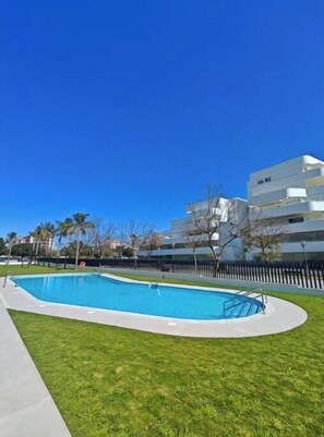 En swimmingpool med en klarblå himmel ovanför, omgiven av en gräsyta och en modern byggnad i bakgrunden.