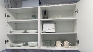Ein weißes Küchenschränkchen mit Regalen, auf denen Teller, eine Kaffeemaschine und Gläser stehen.