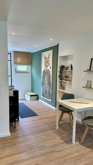 A modern room with a desk, chair, and a large painting of a fox. There is a window with blinds, a shelf with bottles, and a small plant.