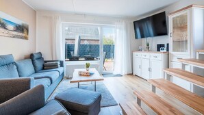 Un salon moderne comprenant un canapé, une table basse et un pouf, un coin cuisine avec des placards et un téléviseur, ainsi qu’une grande fenêtre donnant sur un mur de briques et un espace de détente extérieur.