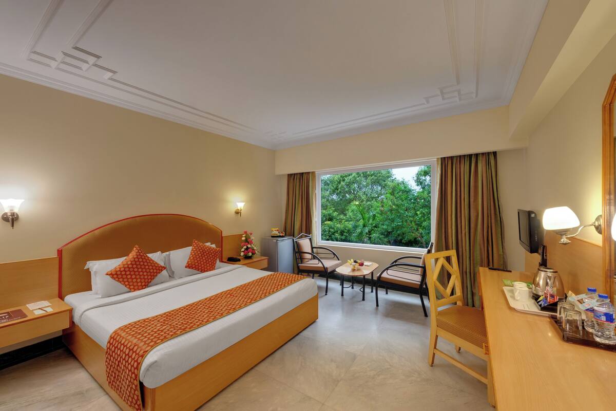 Une chambre d’hôtel comprenant un lit, un bureau, une chaise, une petite table, une fenêtre avec des rideaux et une table de chevet.