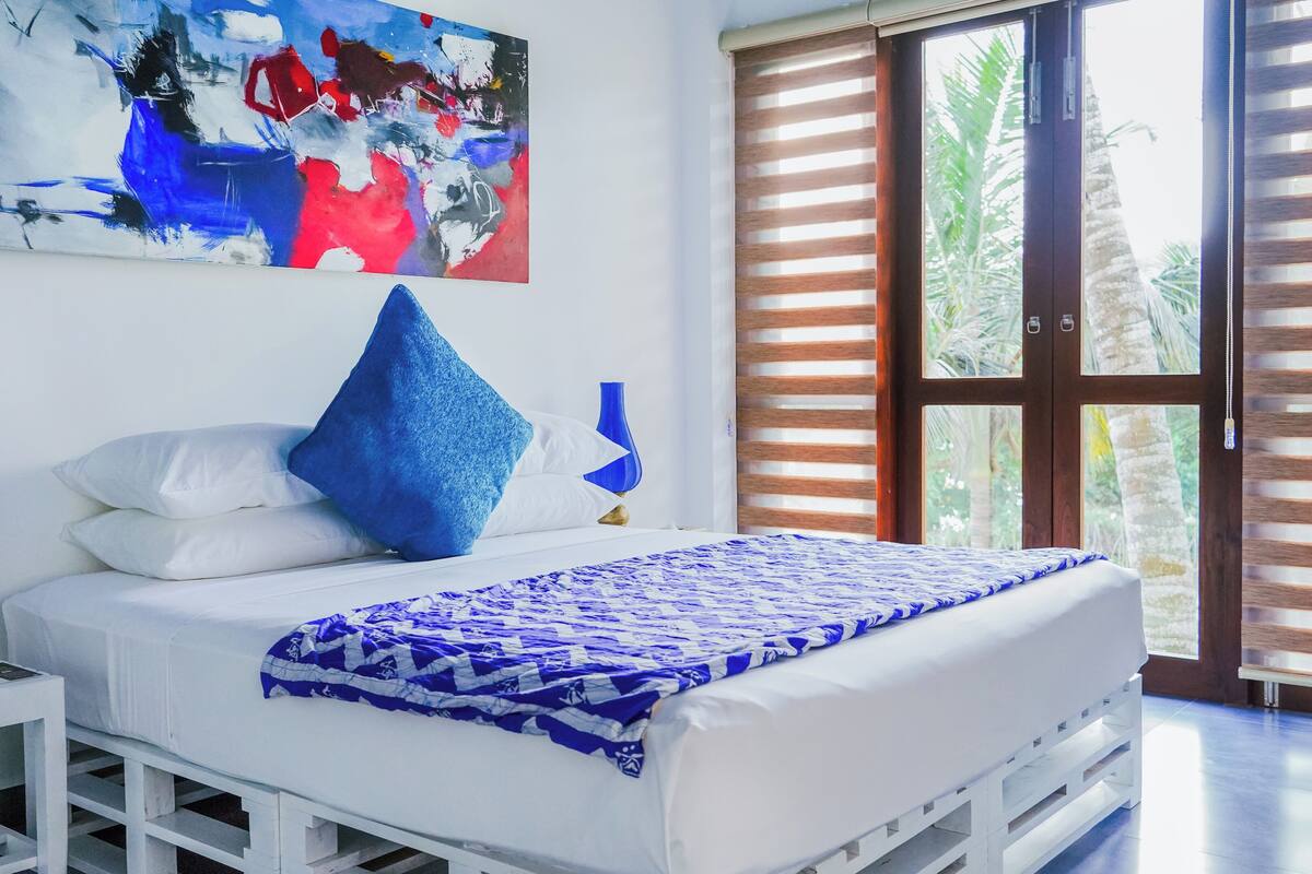 Une chambre à coucher avec un lit, des oreillers, un vase bleu, un tableau et une fenêtre avec des stores.