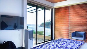 Ein Schlafzimmer mit großem Fenster, einem Bett mit blau-weiß gestreifter Bettdecke, einem Sessel und einem wandmontierten Fernseher.