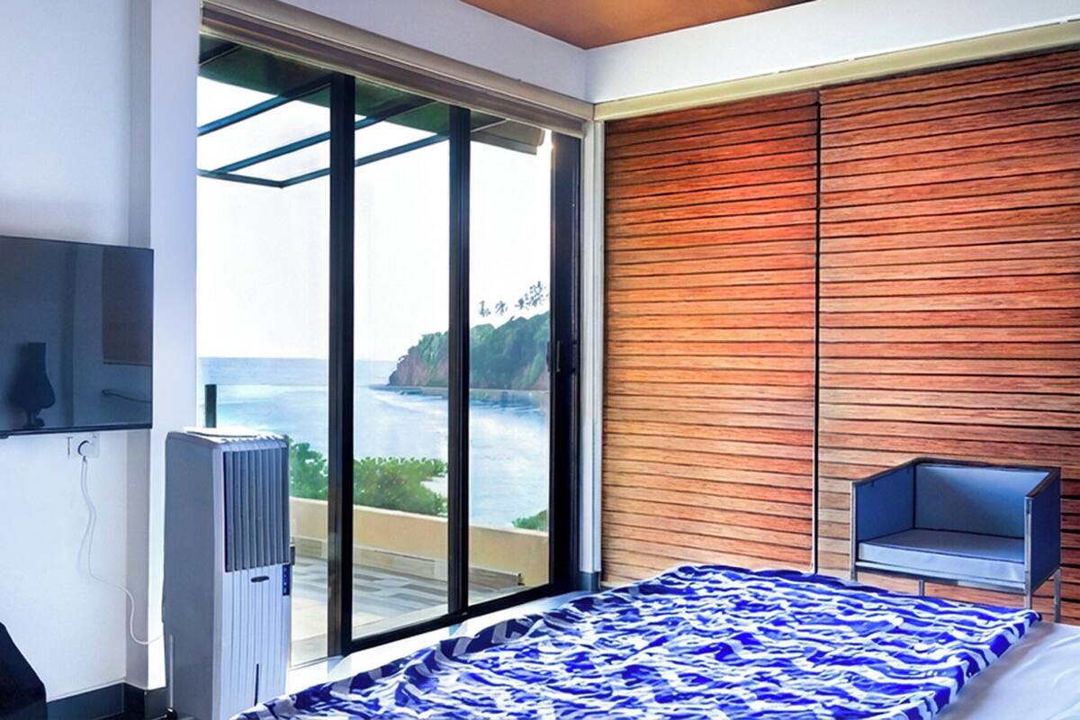 Une chambre avec une grande fenêtre, un lit recouvert d’une couvre-lit à motifs bleus et blancs, une chaise et un téléviseur fixé au mur.