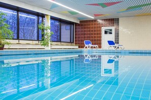 An indoor swimming pool with clear blue water, two lounge chairs, a potted plant, and a view of trees through large windows. - Studio Martina mit Pool und Sauna (Freyung)