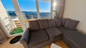 Ein modernes Wohnzimmer mit einer großen Eckcouch, einer Glasschiebetür, die auf einen Balkon mit Bergblick führt, und Holzböden.