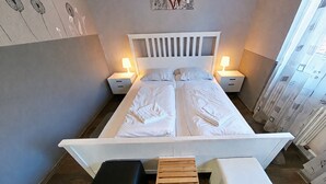Ein Doppelbett mit weißer Bettwäsche, zwei Nachttische mit Lampen, eine Holzbank und eine Wand mit einem floralen Kunstwerk.