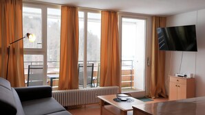 Een hotelkamer met een bank, een televisie, een tafel met een schaal, een radiator en grote ramen voorzien van gordijnen.